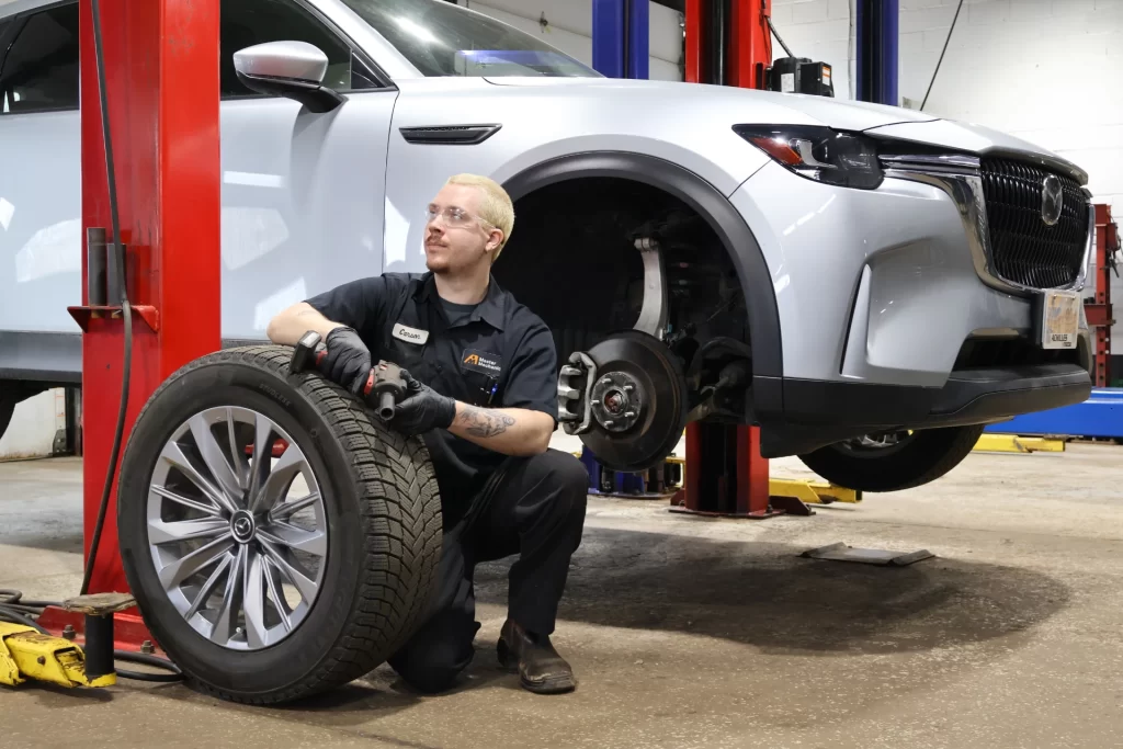A Master Mechanic technician kneels beside a raised silver SUV in an auto shop, holding a power tool and a removed tire.